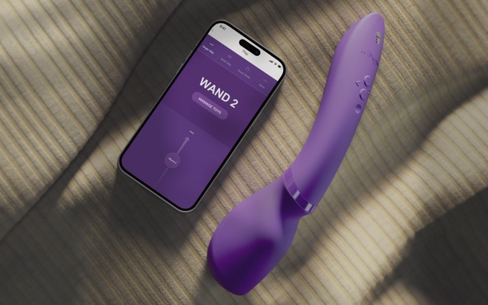 We-Vibe Wand 2 紫色按摩棒與手機並置於織紋布面,螢幕顯示 We-Vibe App 控制介面,自然光影